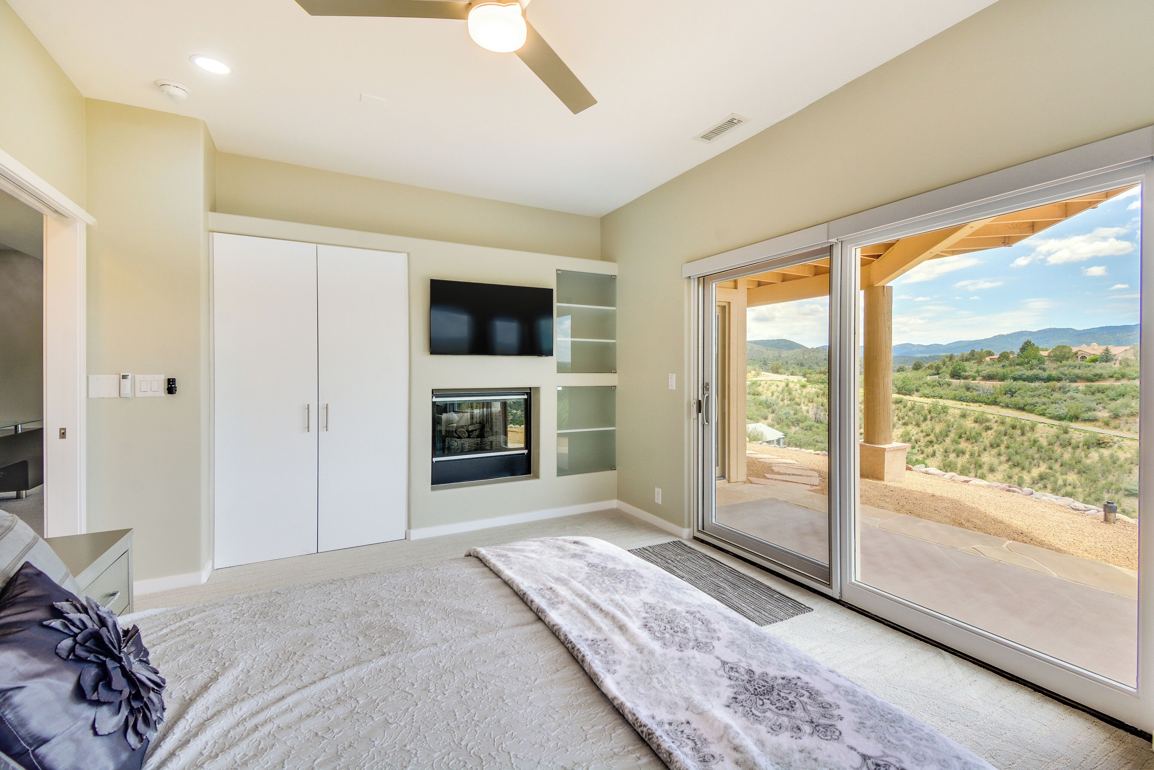 A bedroom with a bed, a ceiling fan, a TV, and a sliding glass door to a balcony with a view of the countryside.