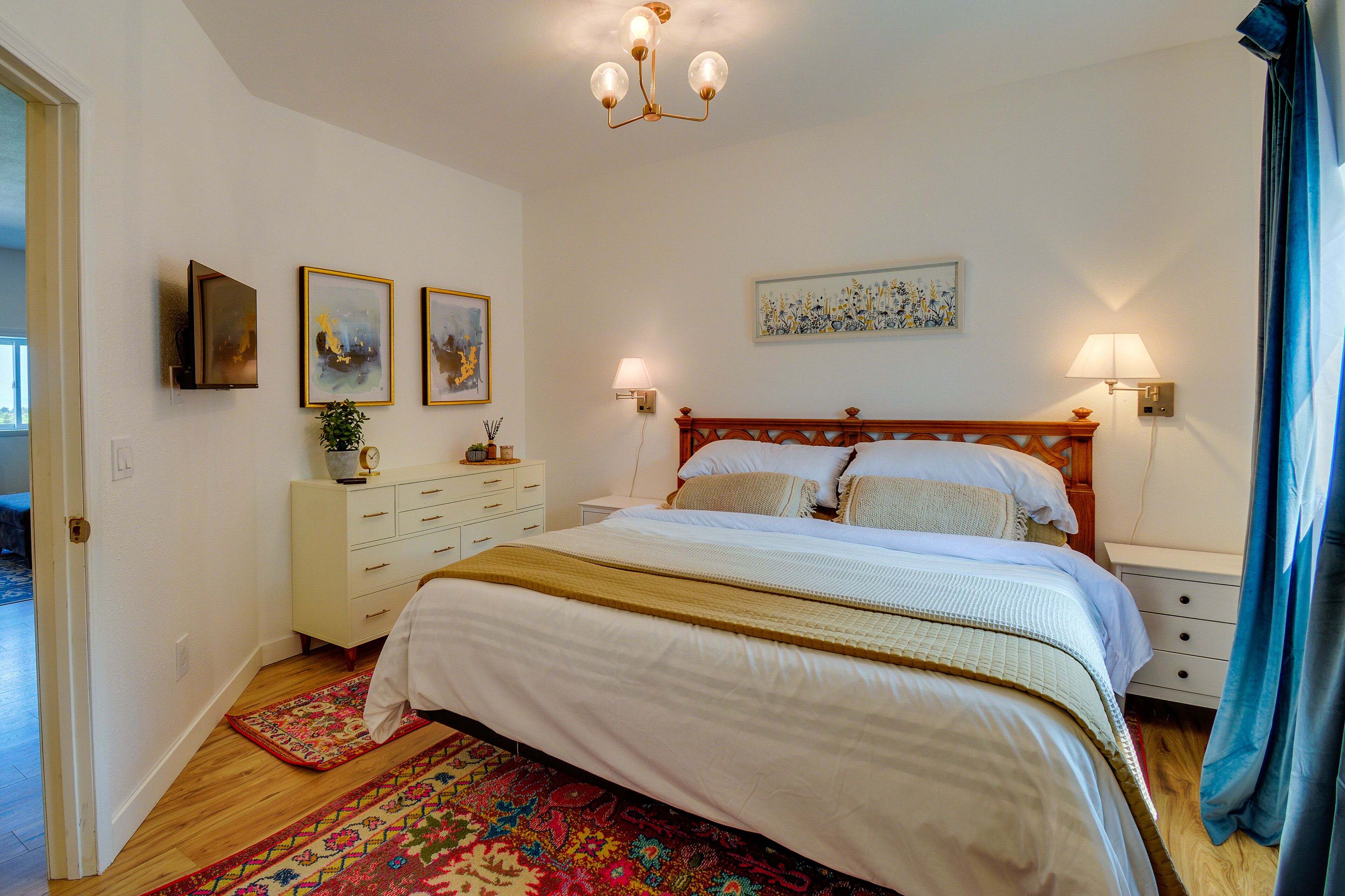 A bedroom with a wooden bed, white bedding, a patterned rug, a nightstand, a wall-mounted lamp, and artwork.