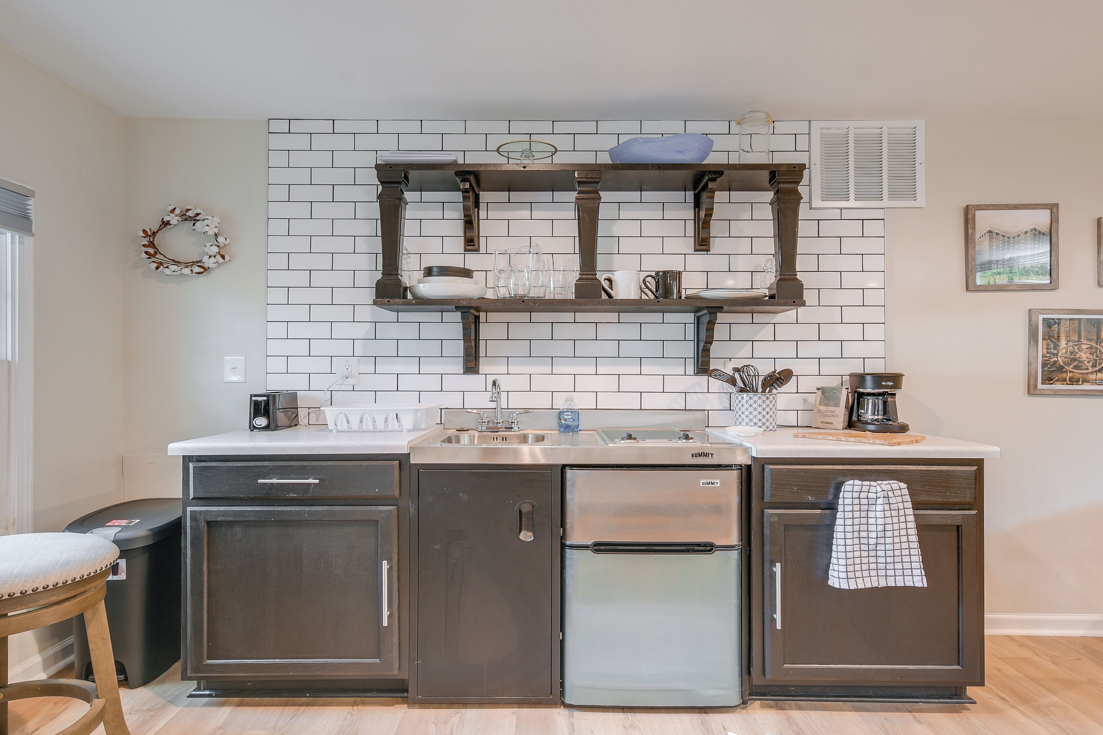 A kitchen with dark cabinets, a stainless steel refrigerator, a white tiled backsplash, and a wooden stool.