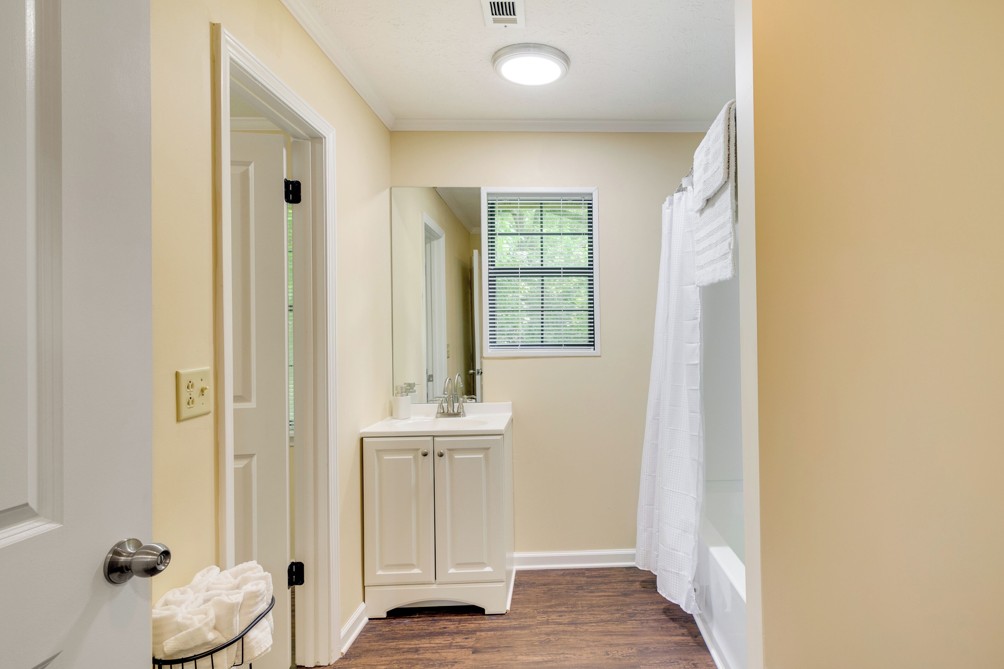 A narrow hallway with a white door, a white vanity with a mirror, and a window with blinds.