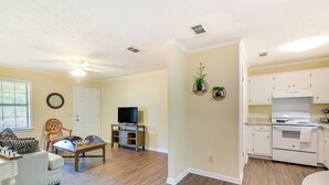 Una sala de estar con un sofá, una mesa de centro y un mueble para la televisión. Hay una cocina con gabinetes blancos y una estufa.