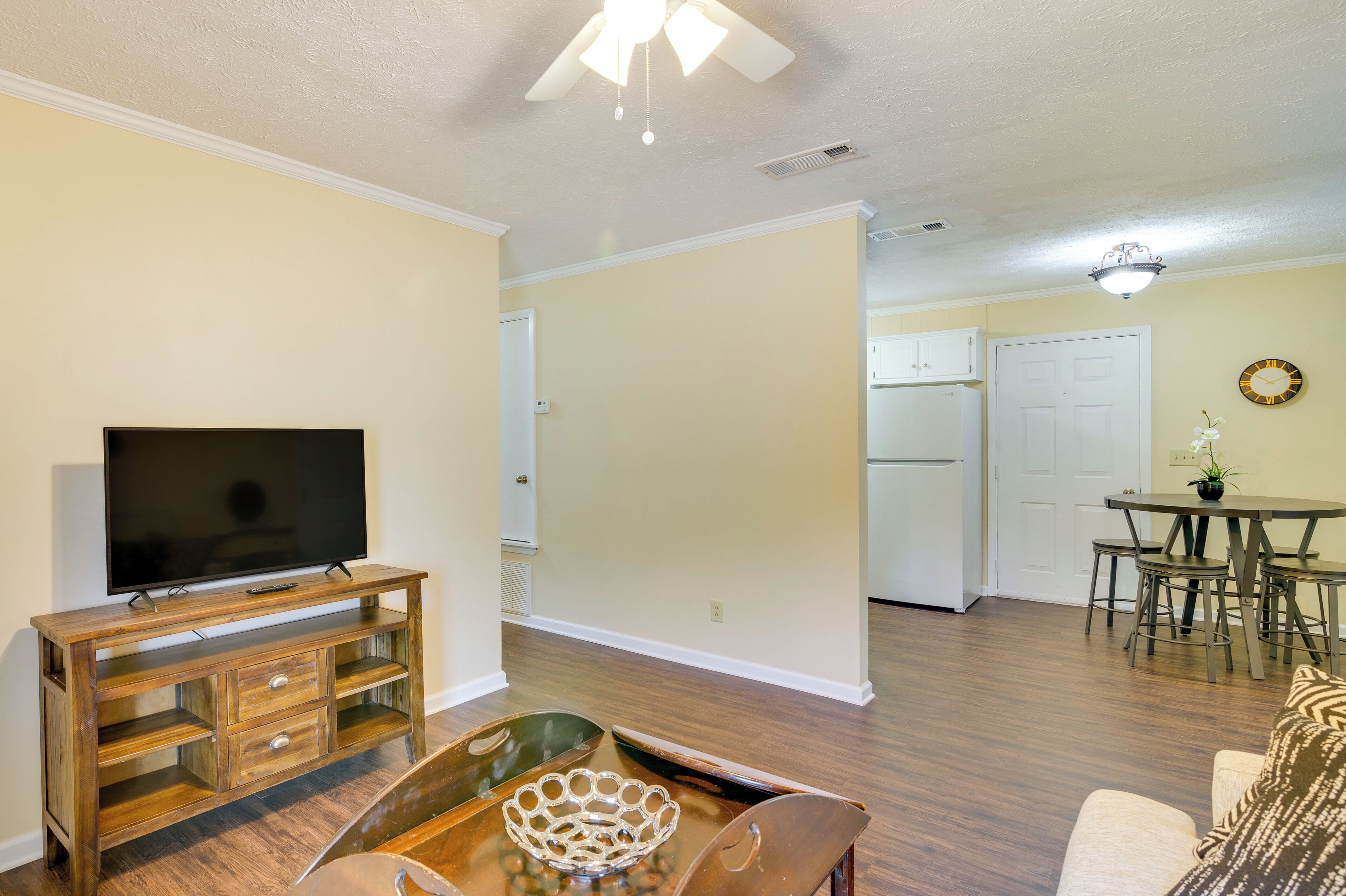 A living room with a TV on a wooden stand, a dining table with chairs, and a kitchen in the background.