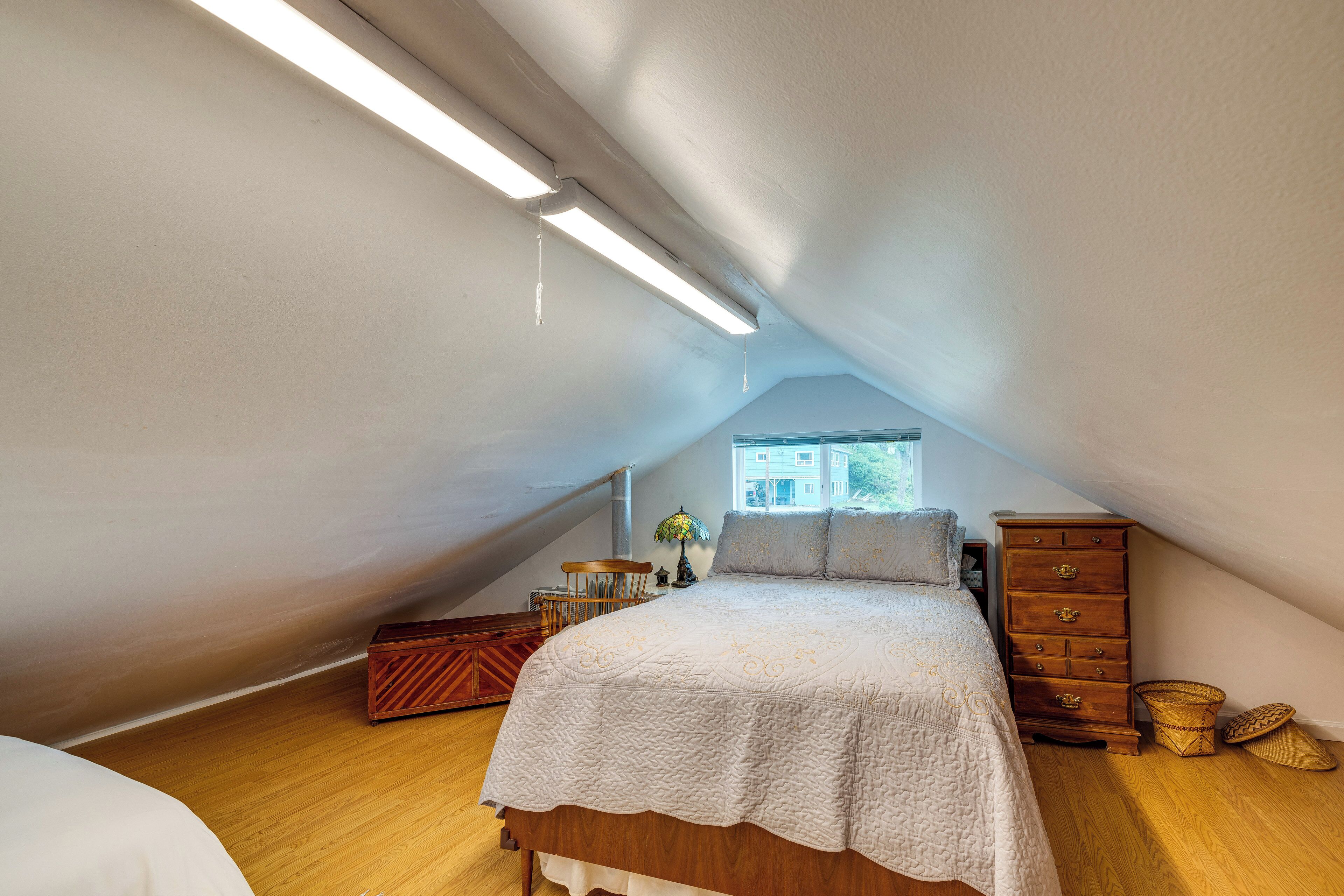 Un dormitorio con techo inclinado, una cama con colcha blanca, un aparador de madera y una ventana con vistas a edificios.