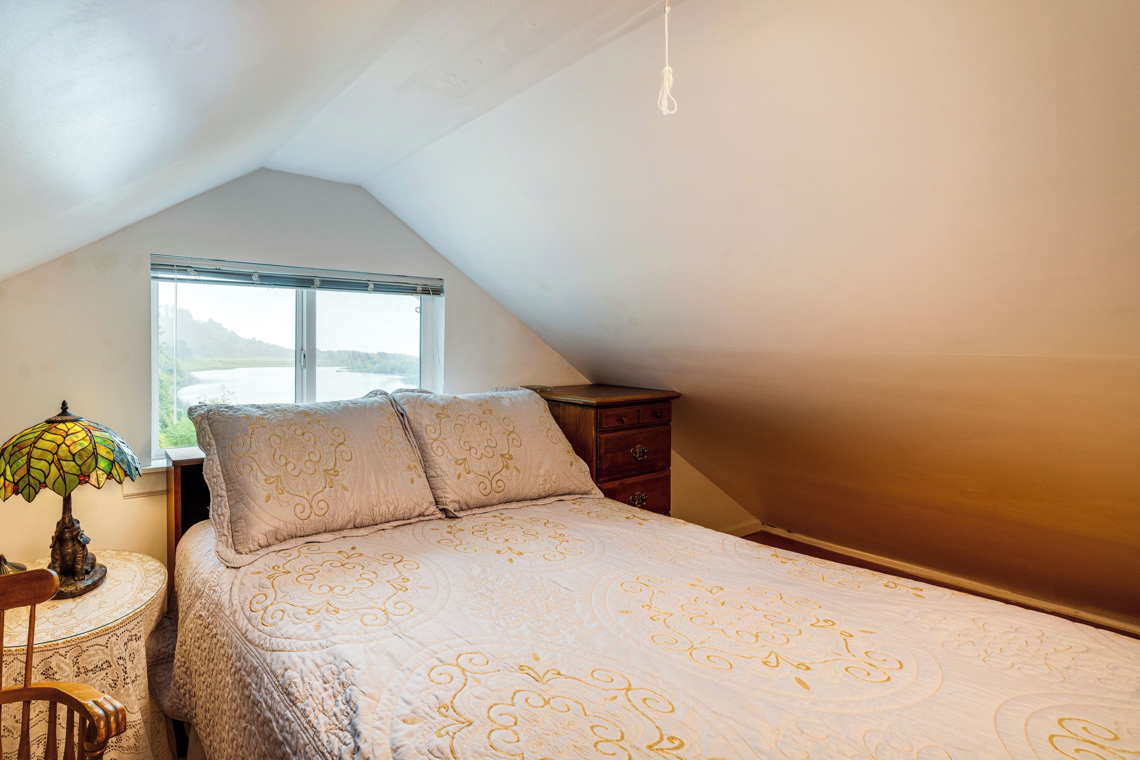 A bedroom with a sloped ceiling, a bed with a patterned bedspread, a nightstand with a lamp, and a window with a view of the outdoors.