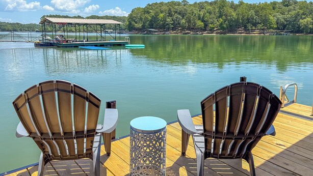 Une terrasse en bois avec deux fauteuils Adirondack et une table en métal, donnant sur un lac paisible avec un quai et une zone abritée en arrière-plan.