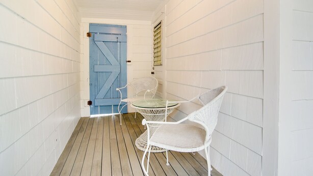 Un couloir étroit aux murs blancs et au plancher en bois, qui mène à une porte bleue. On y trouve une table en verre et des chaises en osier blanc.