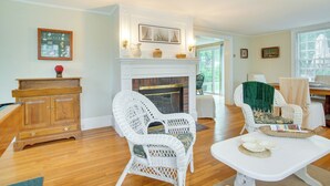 Un salon avec une cheminée, des fauteuils en osier blanc, un buffet en bois et une table à manger.