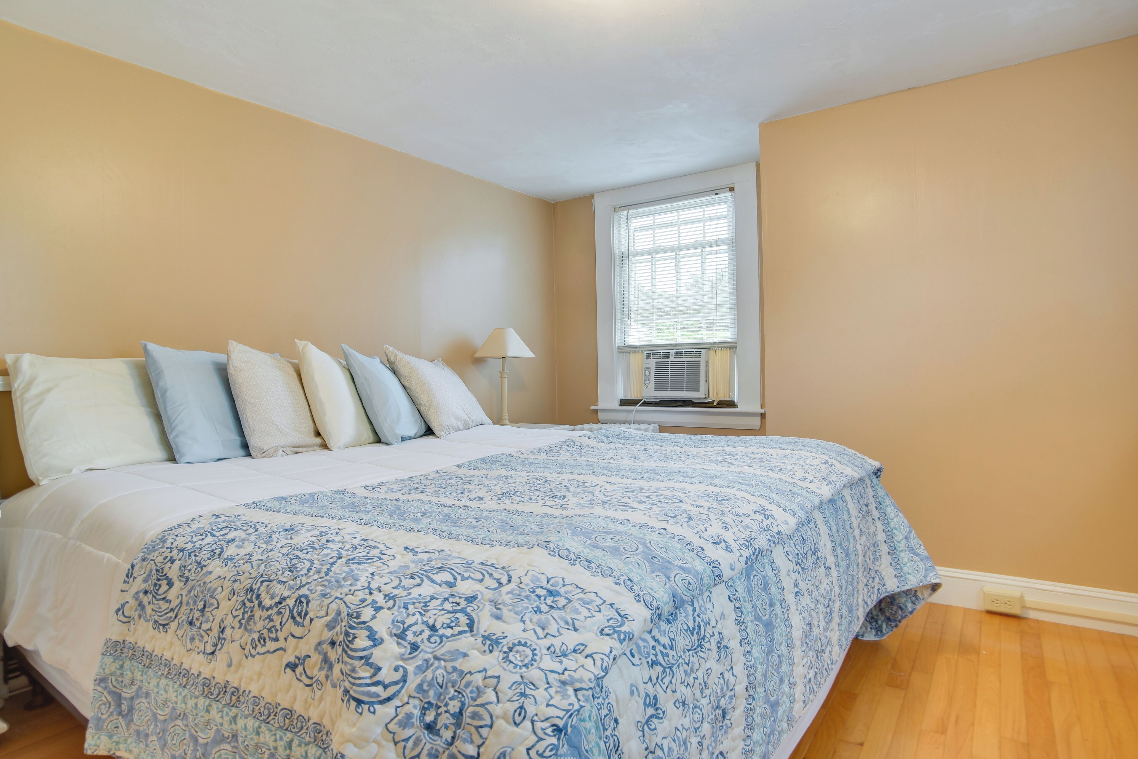 Ein Schlafzimmer mit einem großen Bett, beige Wänden, einem Fenster mit Jalousien und einem Deckenventilator.