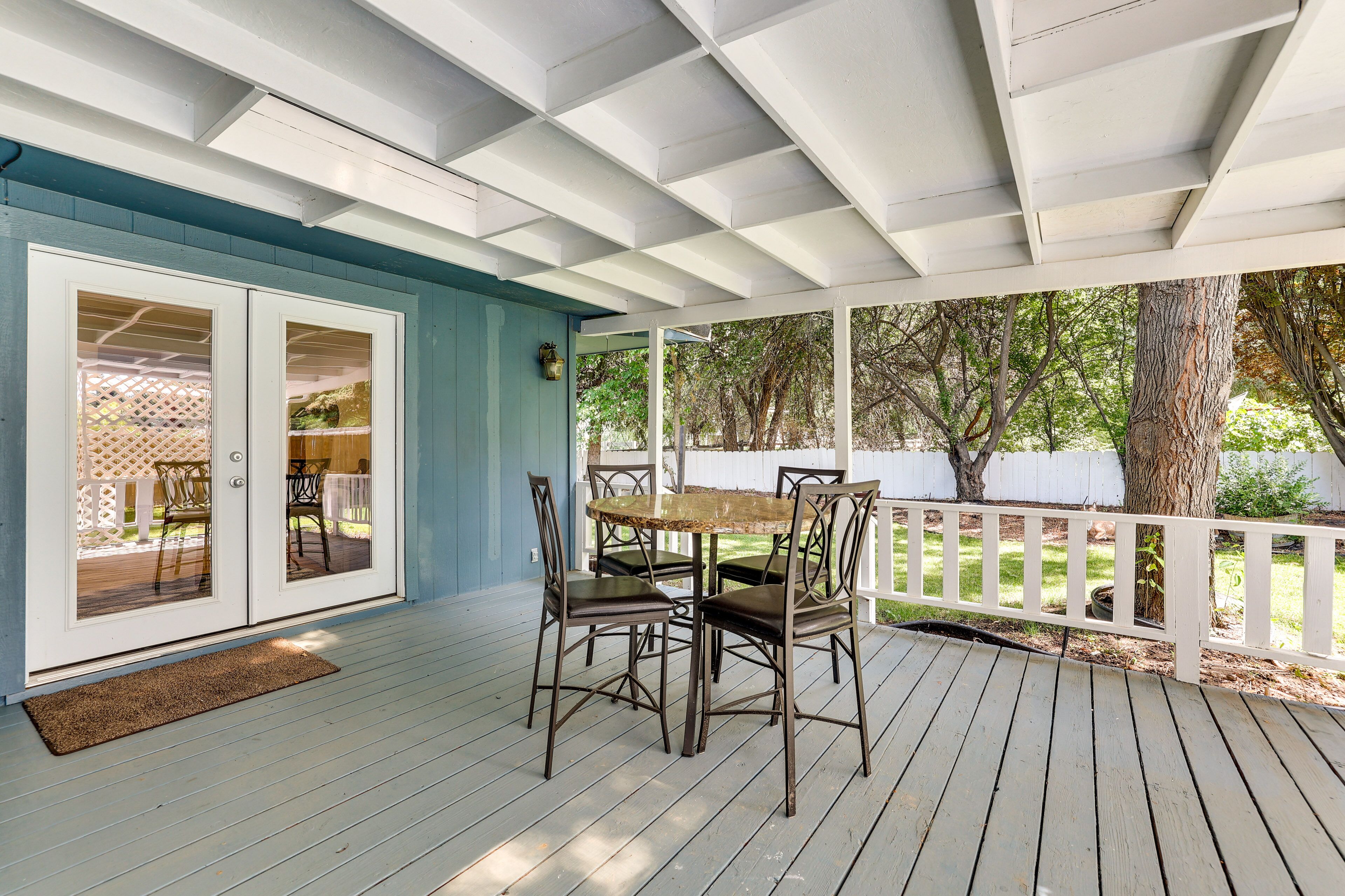 A covered patio with a table and chairs, a white fence, and trees in the background.