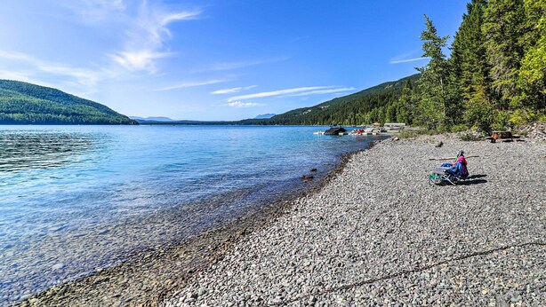 Une plage de galets bordée d’une eau cristalline, une rangée de cabanes et une colline boisée.