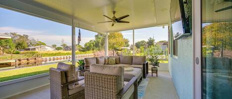 A covered outdoor seating area with wicker furniture, a ceiling fan, and a view of a park and waterway.