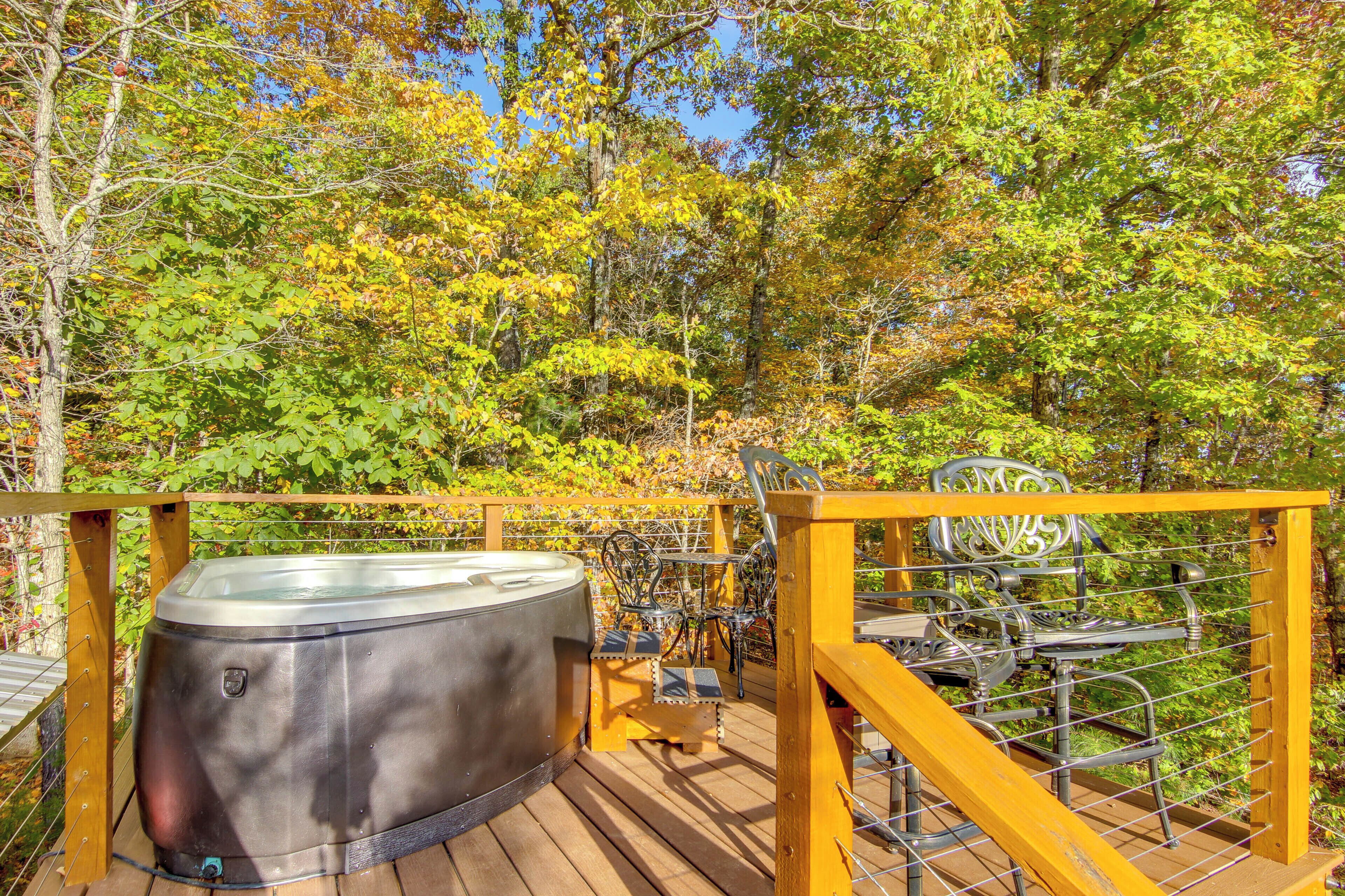 Un bain à remous sur une terrasse en bois, avec des chaises en métal et une rambarde, le tout entouré d’arbres.