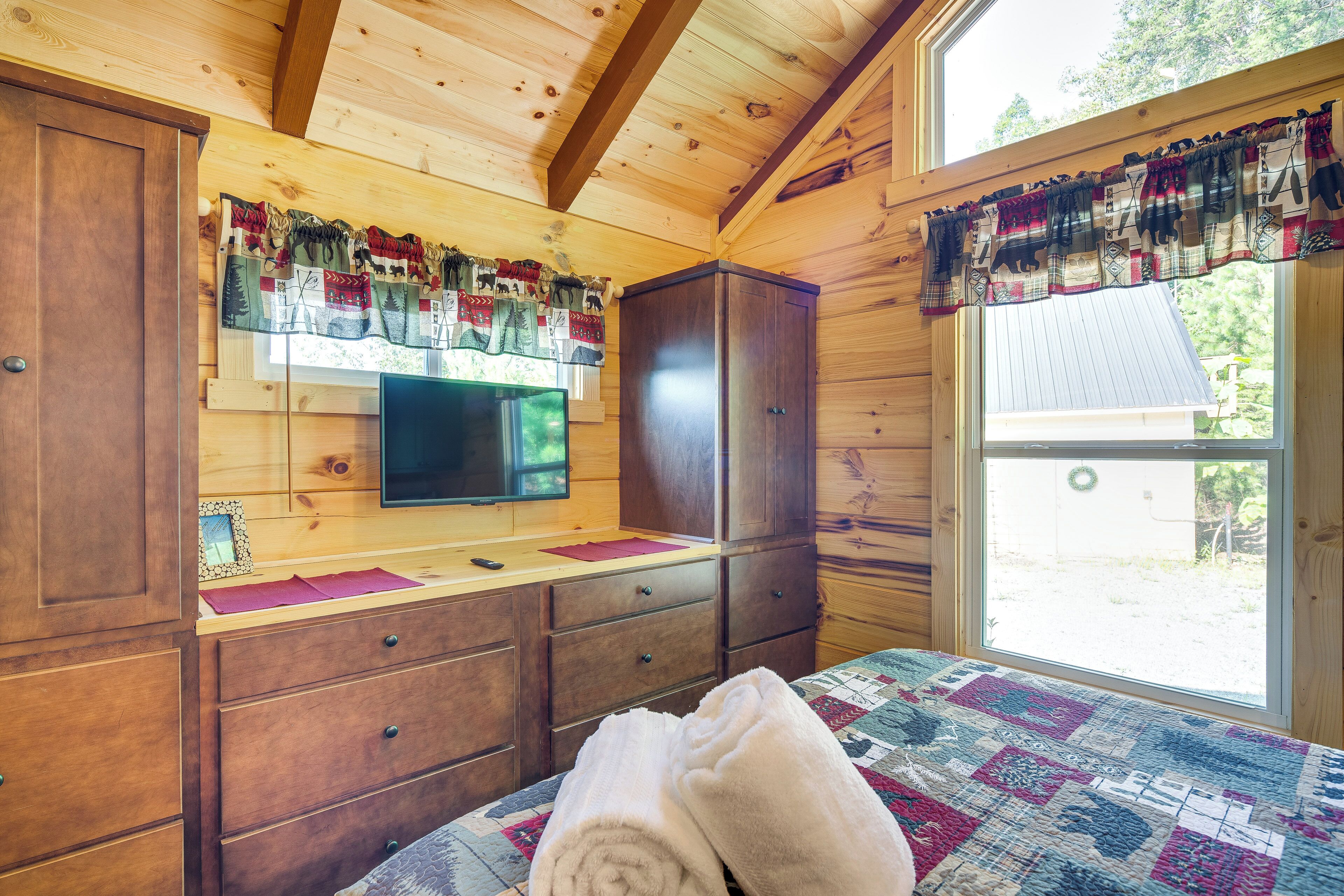 A wooden cabin room with a bed, a TV on a wooden shelf, and a window with curtains.