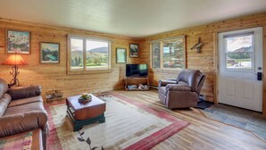 A cozy living room with wooden walls, a flat-screen TV, leather sofas, and a large window offering a scenic view.