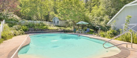 A swimming pool with a wooden deck, surrounded by a white fence and lush greenery.