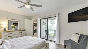 Une chambre à coucher comprenant un lit, une commode, une chaise, un ventilateur de plafond, un téléviseur et une porte-fenêtre coulissante donnant sur un balcon.
