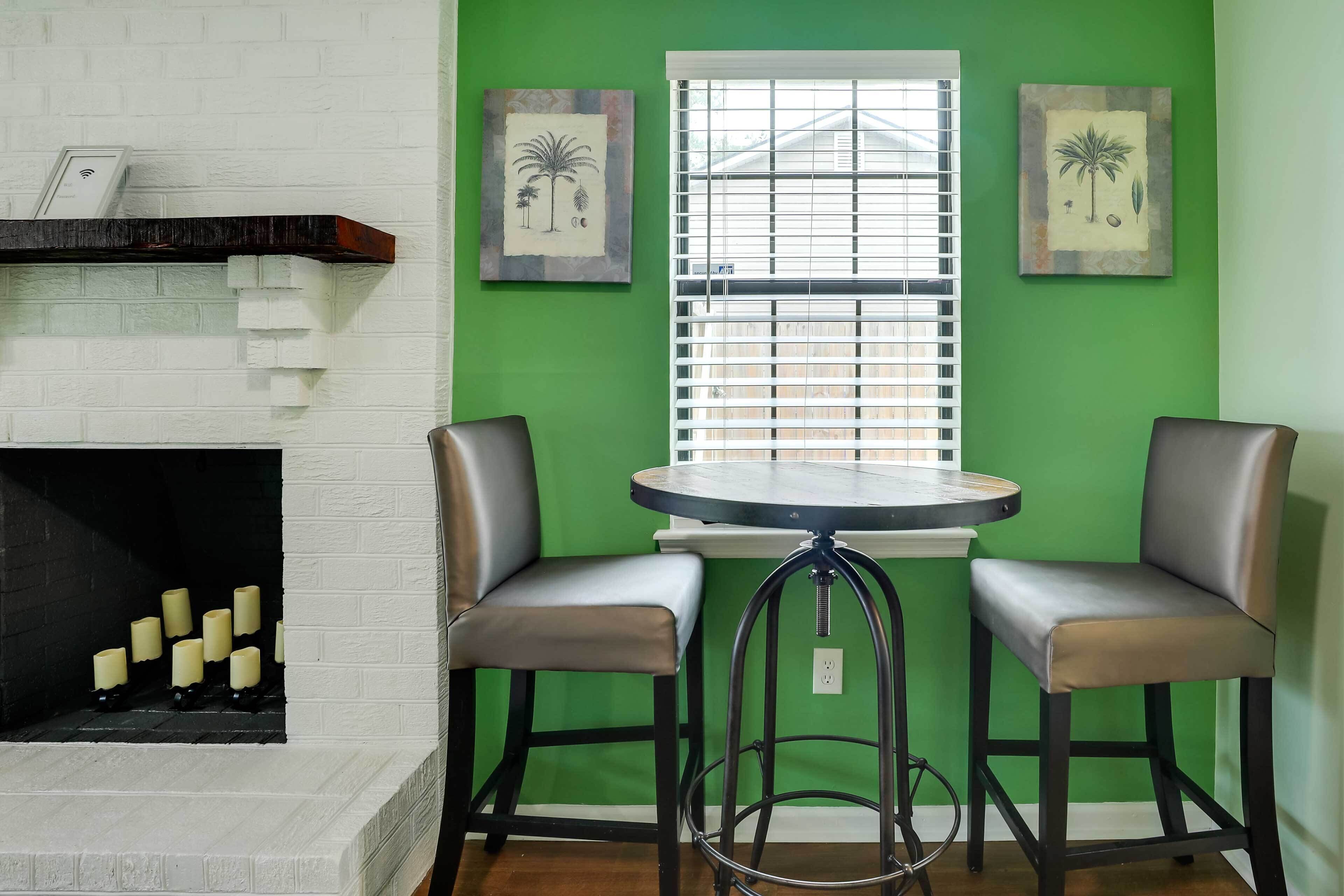 A room with a white brick fireplace, green walls, two bar stools, a round table, and two framed artworks.