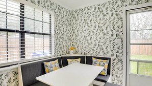 A corner seating area with a white table and grey cushions, a window with blinds, and a door leading outside.