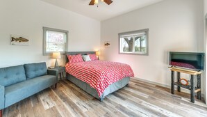 Una camera da letto con un letto, un divano grigio, una ventola a soffitto, un televisore su un mobile in legno e una finestra con vista sugli alberi.