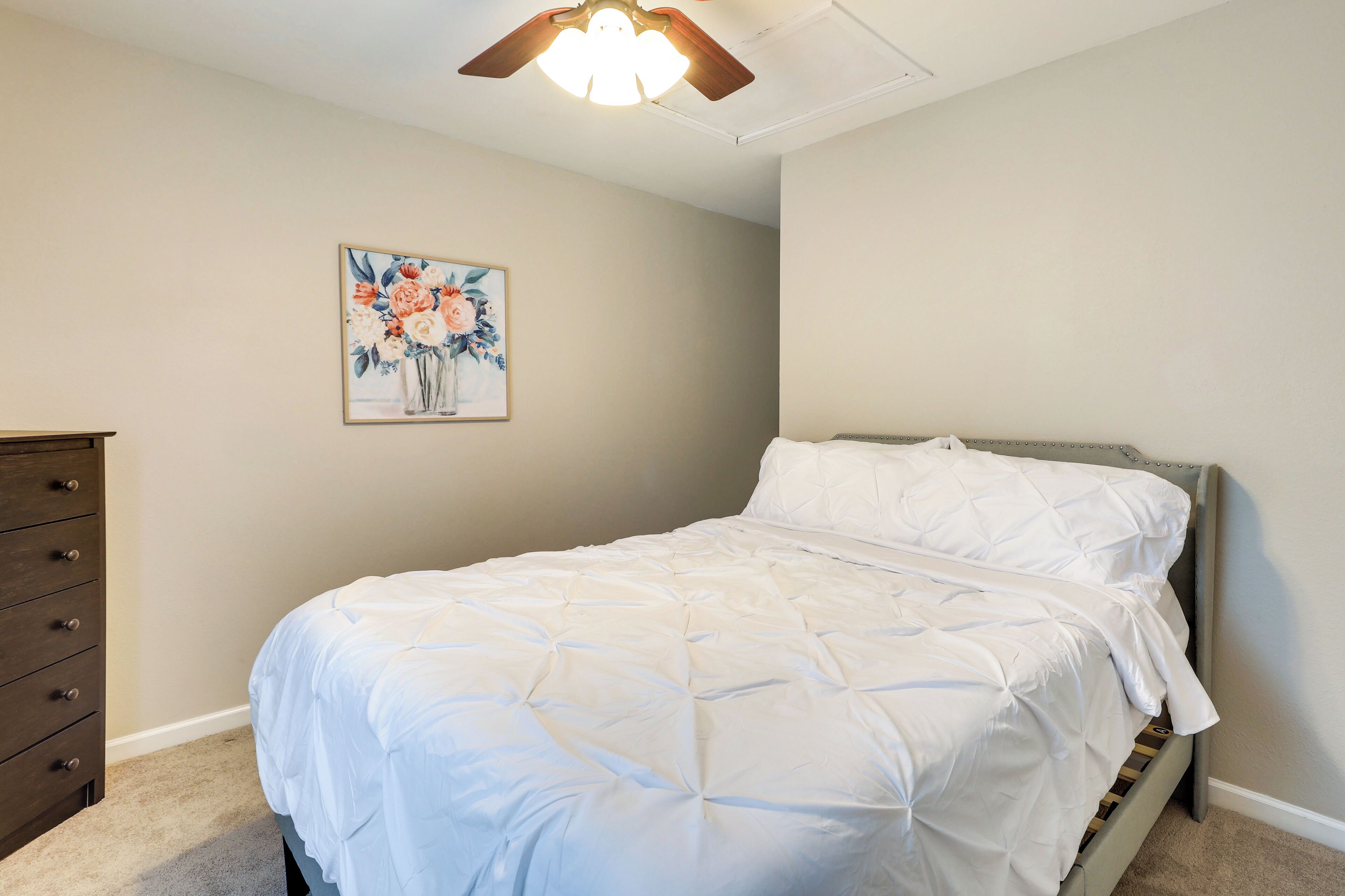 A bedroom with a bed, a dresser, a ceiling fan, and a floral painting on the wall.