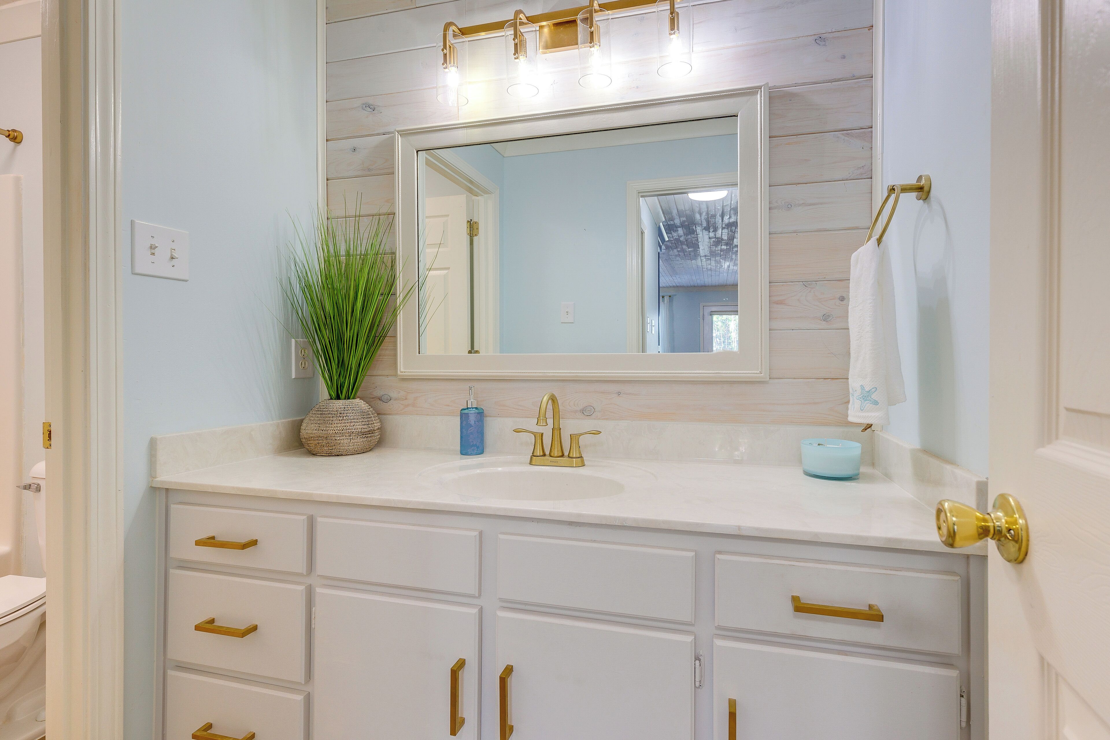 Un baño con tocador blanco, pomos dorados, un espejo grande y un panel de pared de madera.