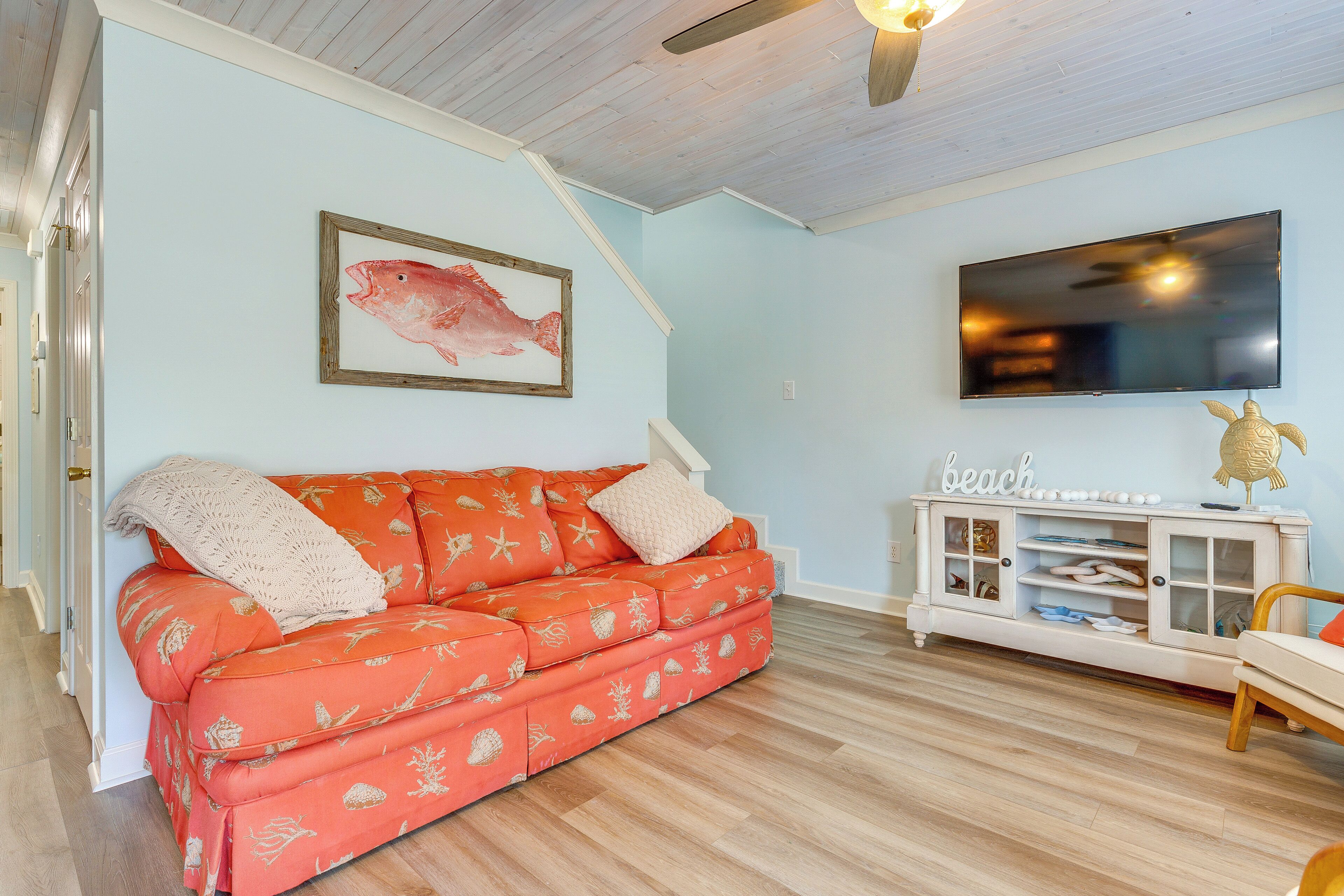 A living room with a light blue wall, a wooden ceiling, a red sofa with a starfish pattern, a white entertainment unit with a 'beach' sign, and a flat-screen TV.