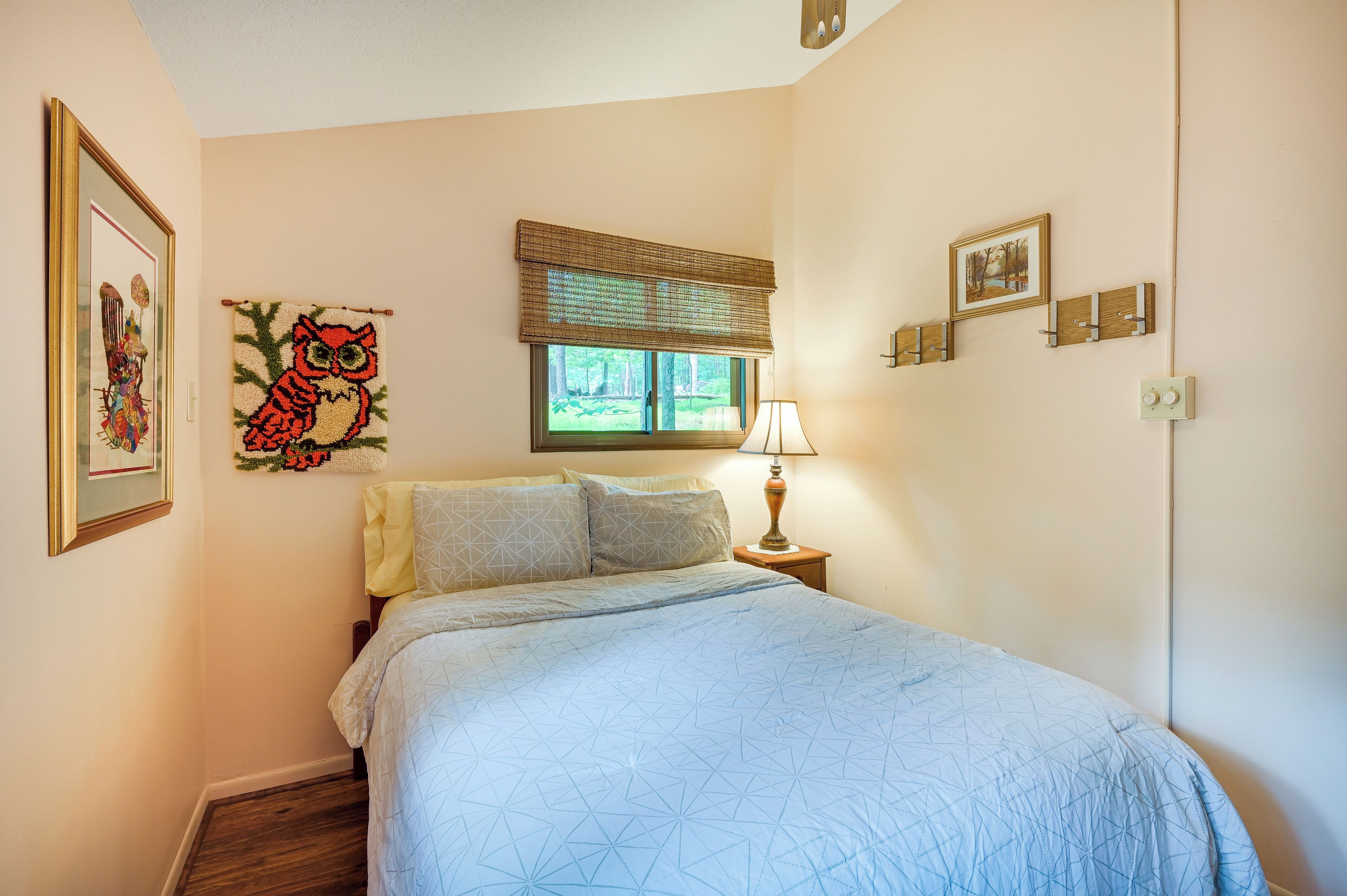 A bedroom with a bed, a window with a blind, a bedside lamp, and wall-mounted picture frames.