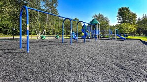 A playground with blue and green play structures, including slides and swings, set on a gravel surface. - Pet-friendly Pocono Lake Cabin w/ Community Pool! (Pocono Lake)