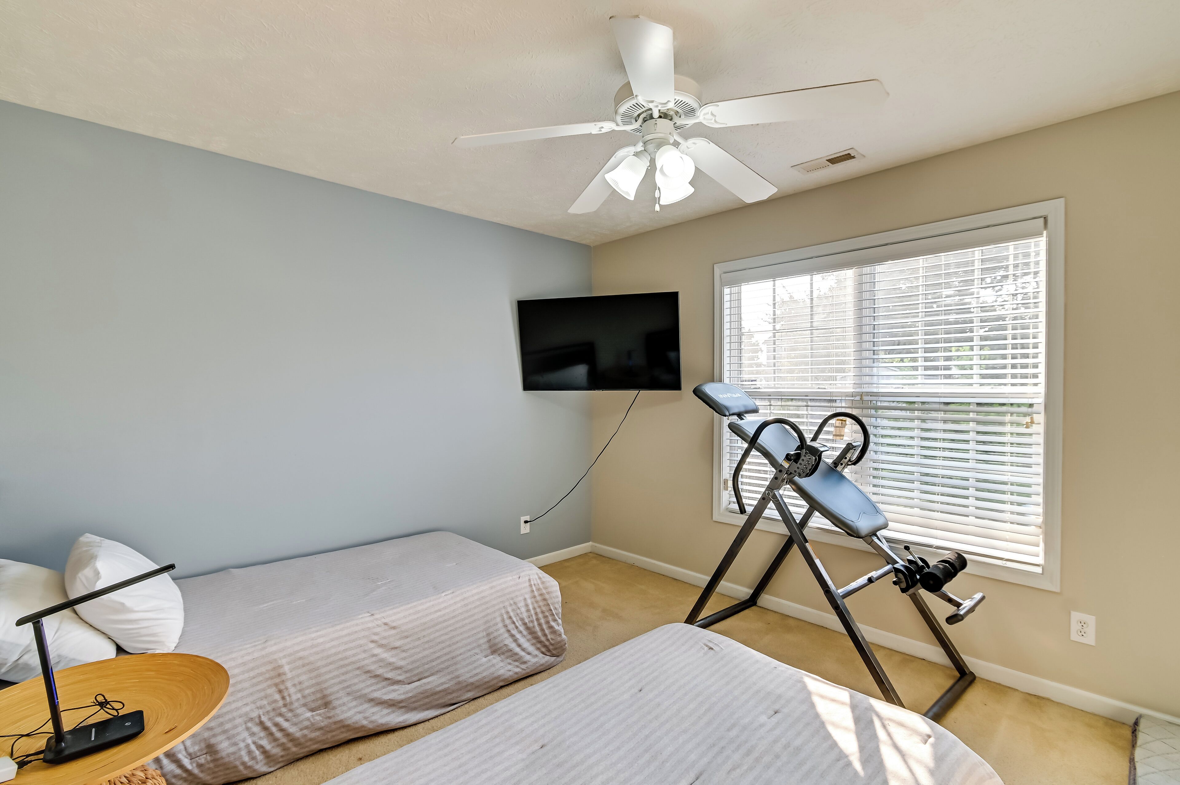 Un dormitorio con dos camas individuales, un ventilador de techo, un televisor montado en la pared y una ventana con persianas.