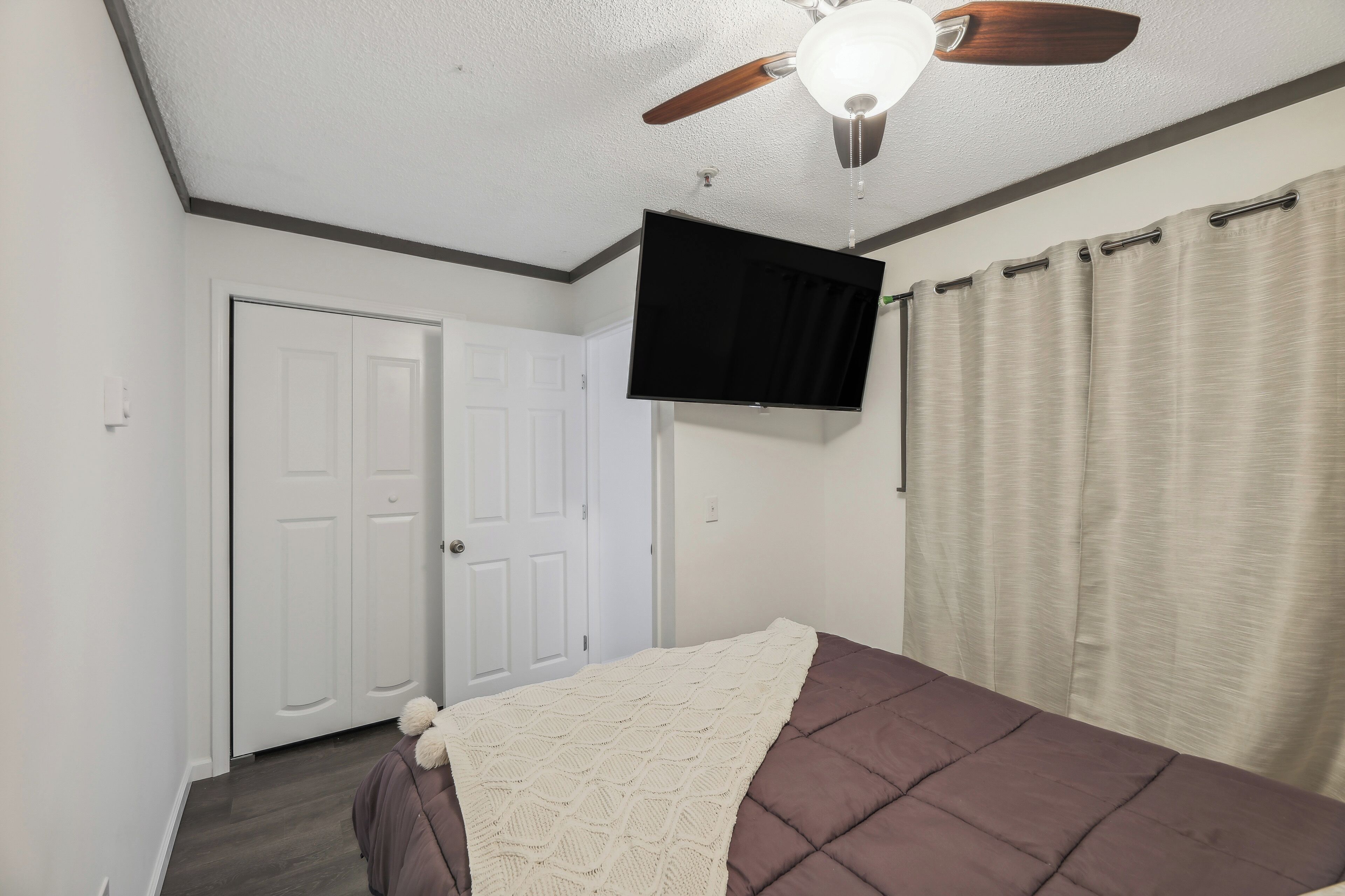 A bedroom with a bed, a ceiling fan, a flat-screen TV, and a door.
