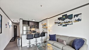 Uma sala de estar moderna com uma mesa de centro de vidro, um sofá cinza com almofadas azuis e uma área de cozinha com um conjunto de banquetas.