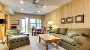 A room with a wooden paneled wall, a ceiling fan, a wooden table, a green sofa, a green armchair, and a dining table with chairs. - Relaxing Saluda Home Near Waterfall Trails! (Saluda)