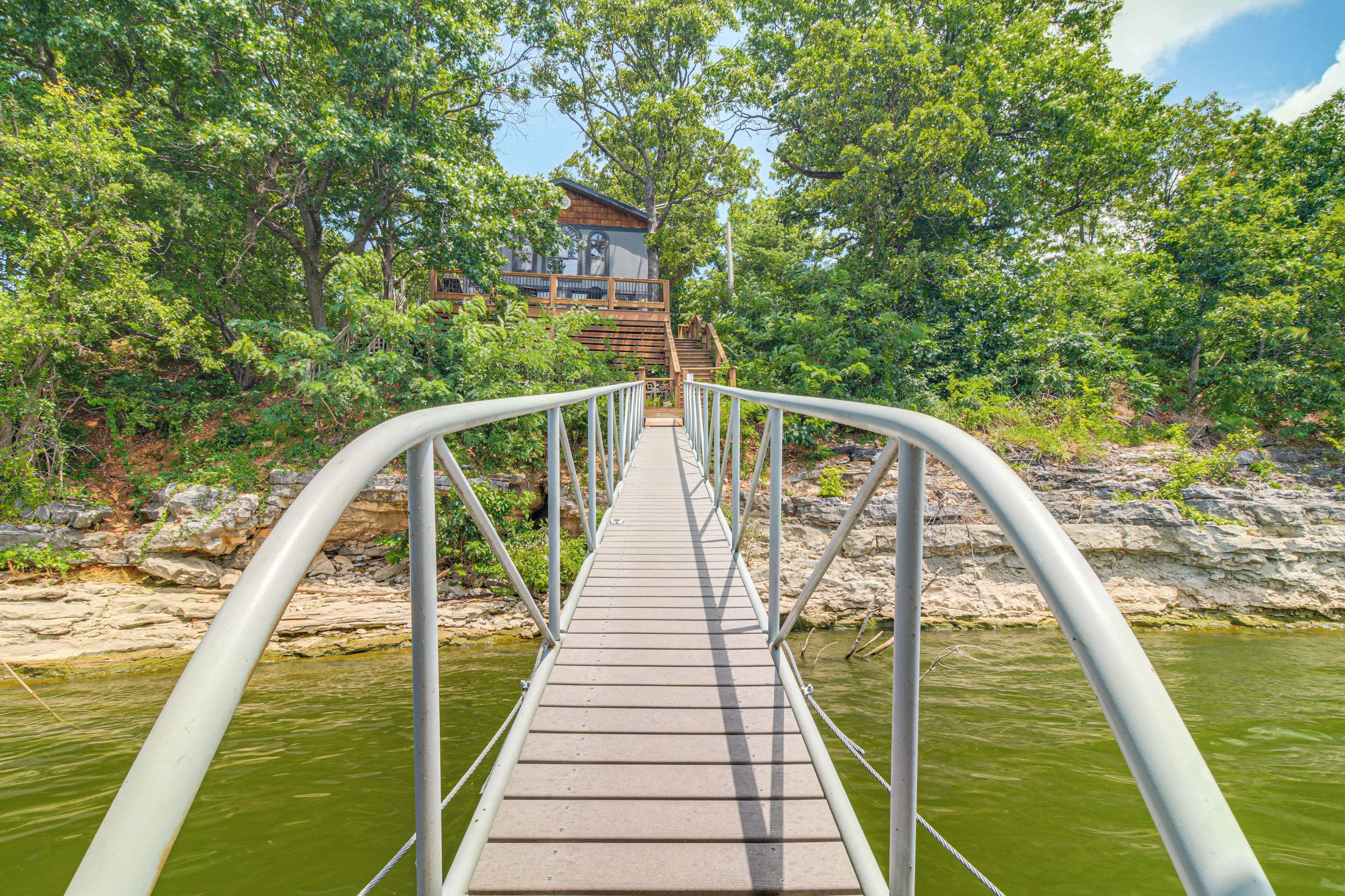 A wooden bridge with metal railings leading to a building surrounded by trees and a body of water.