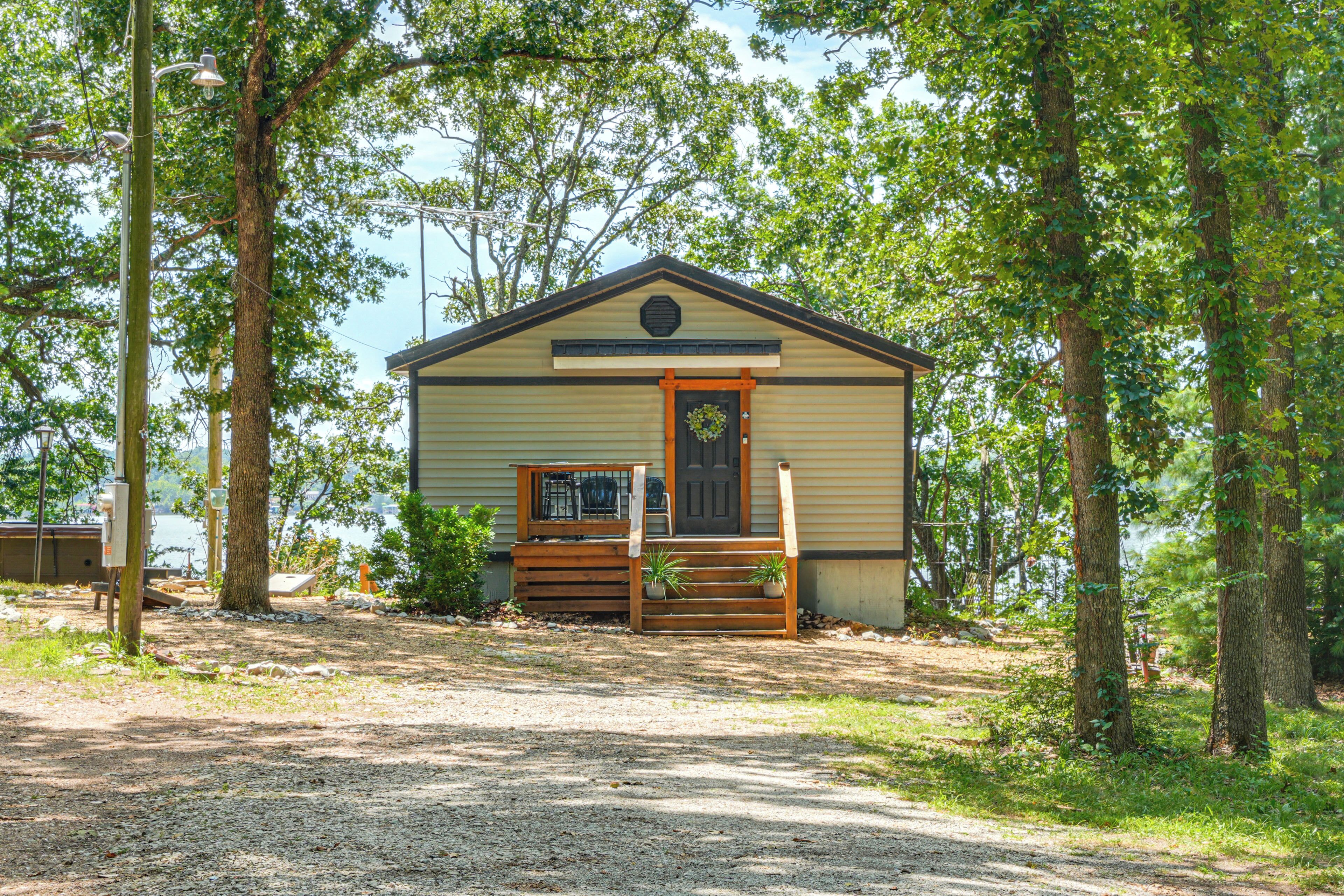 A small, single-story cabin with a front porch, surrounded by trees and a gravel path.