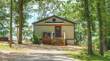 A small, single-story cabin with a front porch, surrounded by trees and a gravel path.