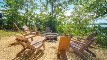 An outdoor seating area with wooden chairs and a central fire pit surrounded by trees.