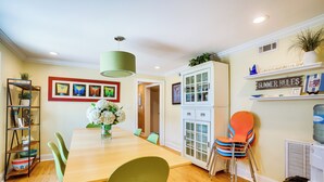 A dining area with a wooden table, green and orange chairs, a hanging light fixture, and a cabinet with glass doors. - Dewey Beach Cottage Near Ocean - Walk to Beach! (Dewey Beach)