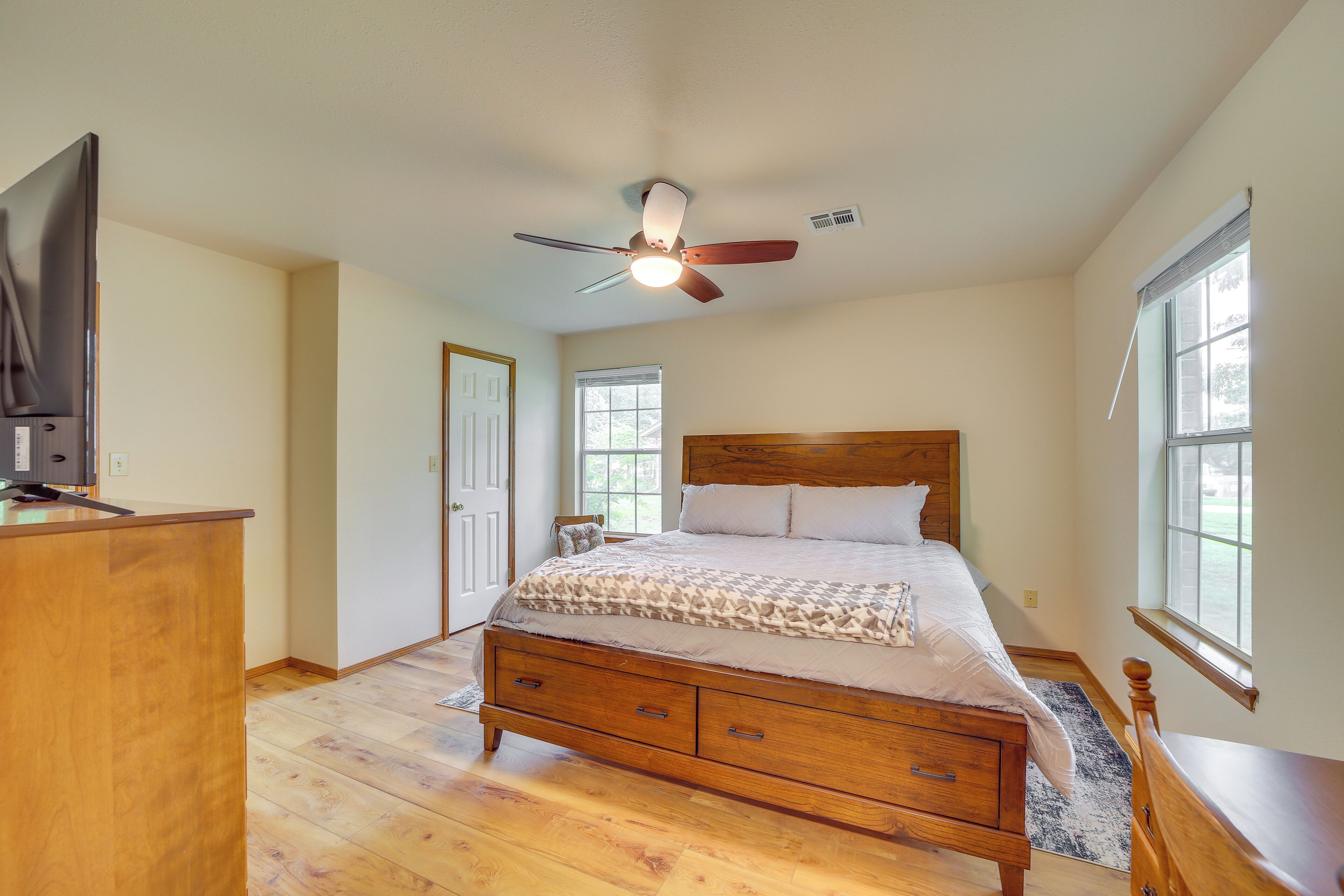A bedroom with a wooden bed, a ceiling fan, a television on a wooden stand, and a window.