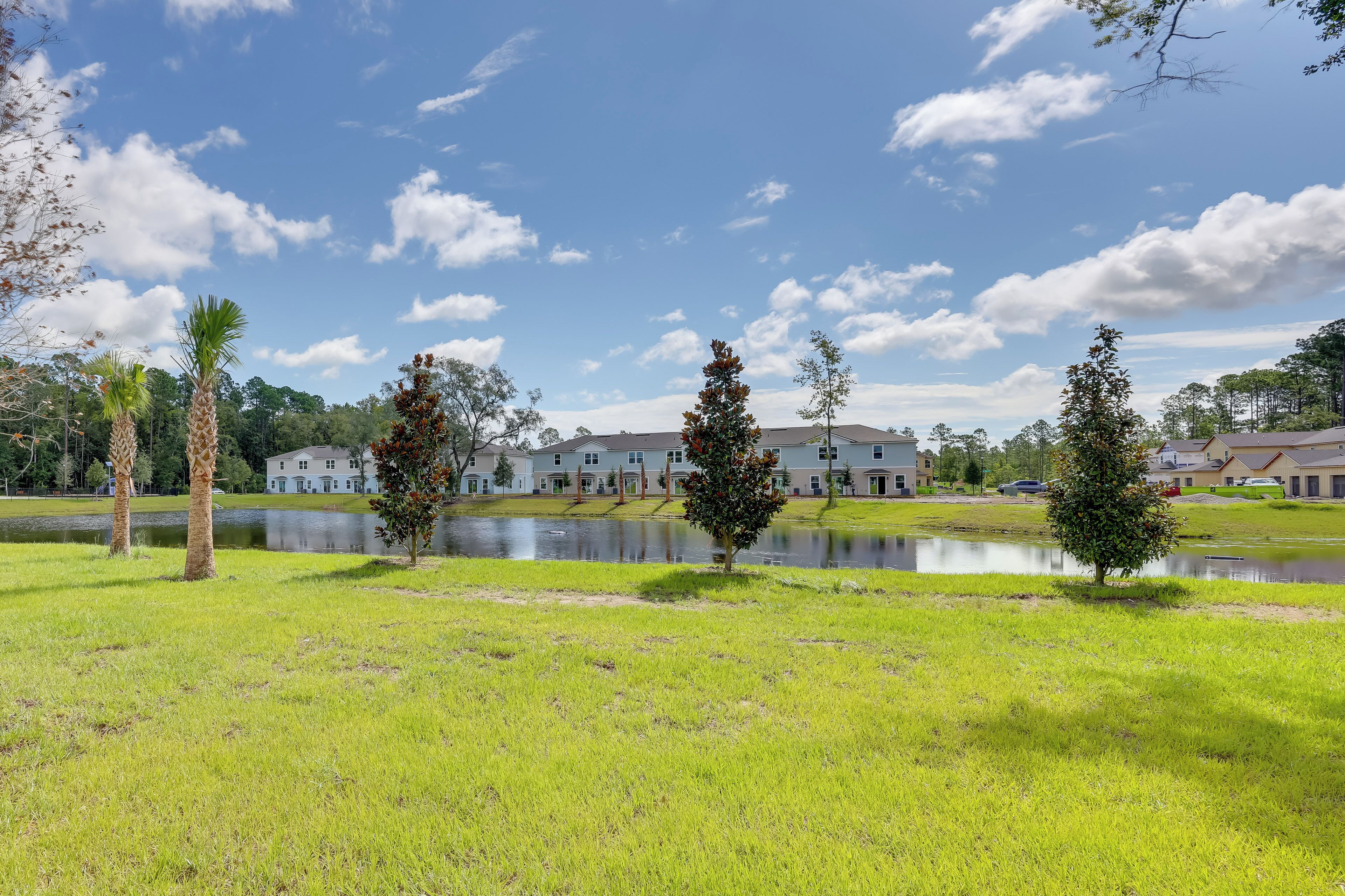 A grassy area with a pond and several trees, with buildings in the background.