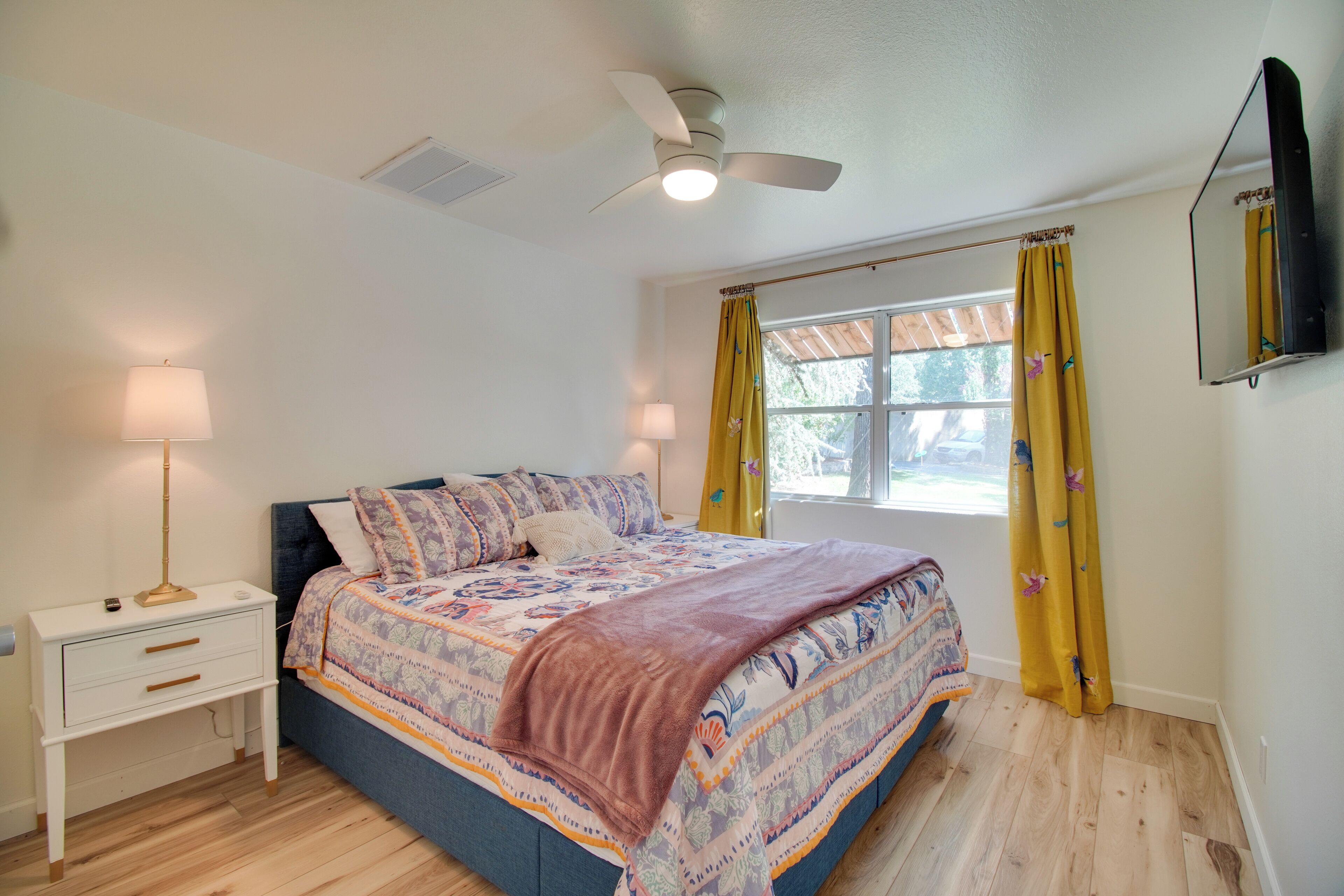 A bedroom with a bed, bedside table, lamp, ceiling fan, window with curtains, and a wall-mounted TV.