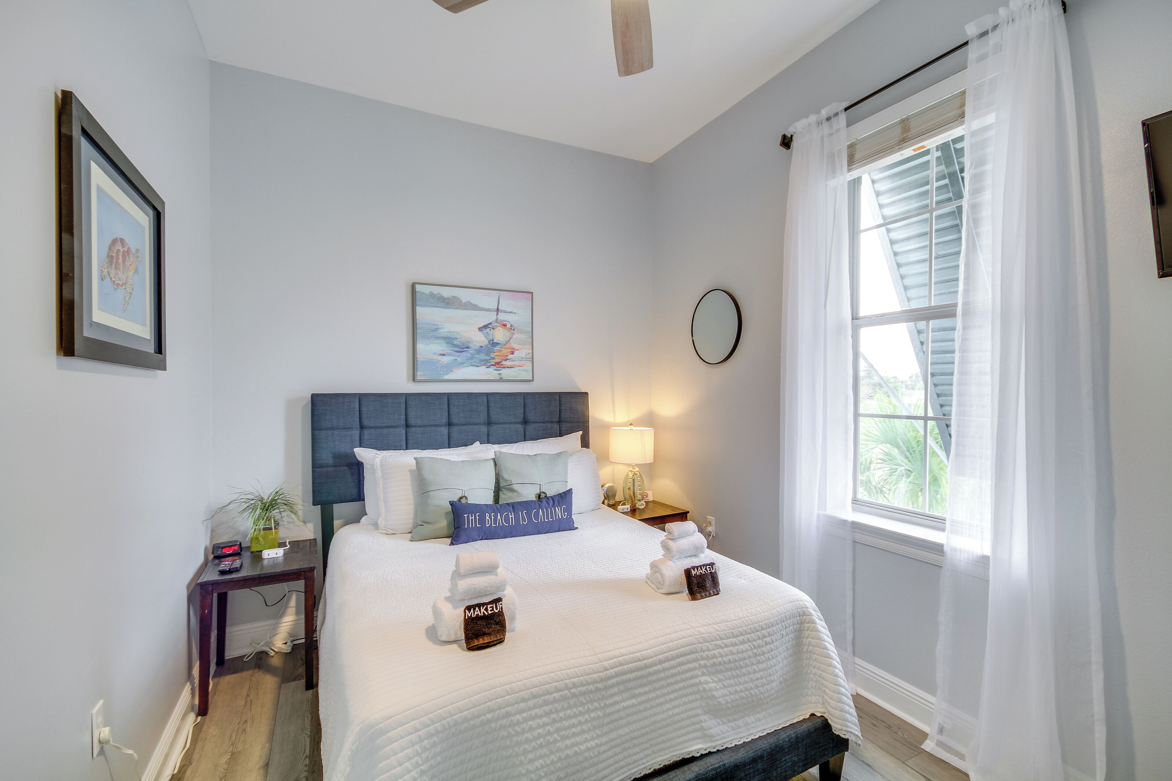 A neatly arranged bedroom with a bed, pillows, a bedside table with a lamp, a framed picture, and a window with sheer curtains.