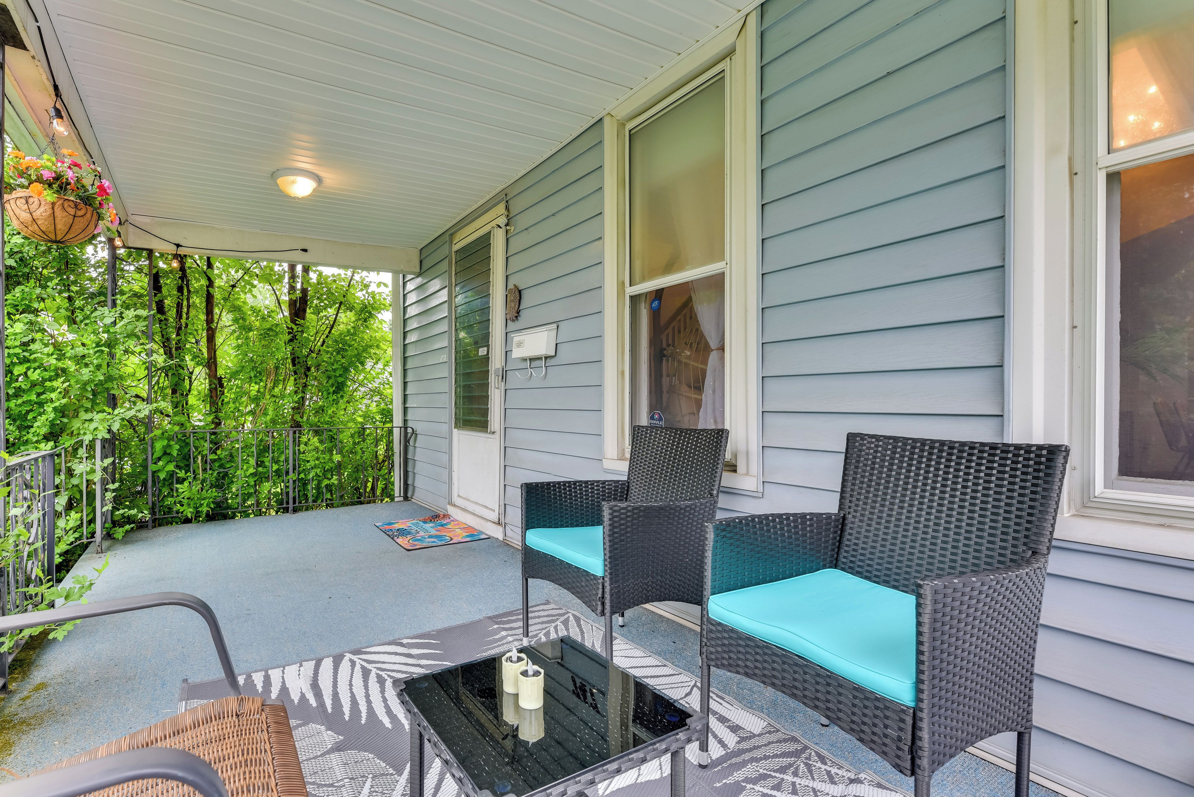 A covered porch with wicker furniture and a glass table, surrounded by greenery.