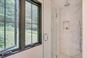 Ein modernes Badezimmer mit Duschkabine aus Glas, einem Fenster mit Blick auf Grünflächen und einer Marmorwand.