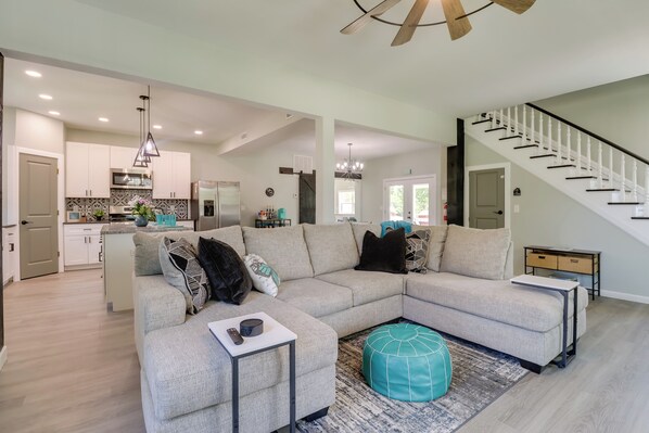 A spacious living area with a large L-shaped sectional sofa, a small side table, and a teal ottoman. A staircase with white balusters leads to an upper level. - Spacious Yard, Games & Fire Pit: Grove Home Rental (Grove)