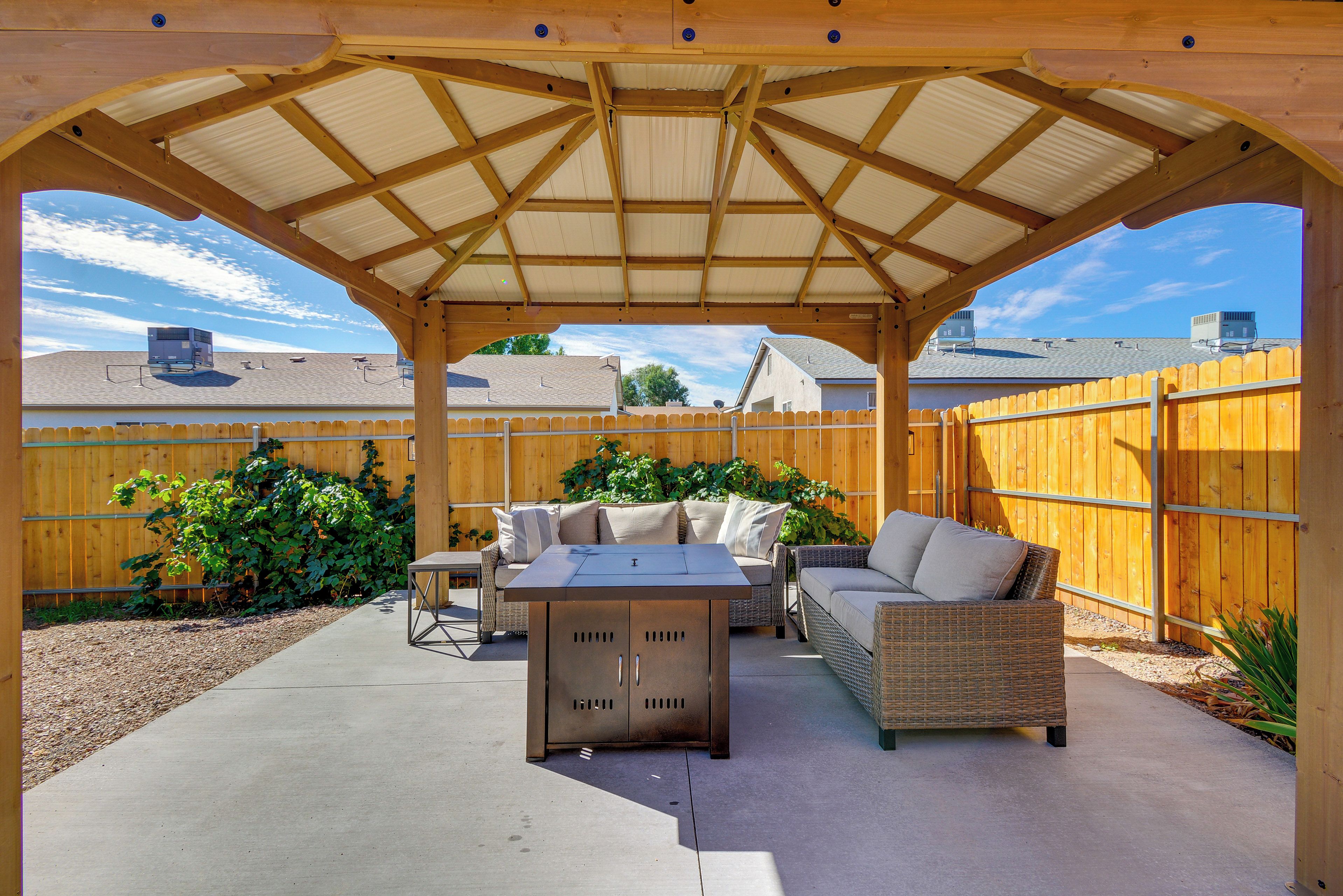Une terrasse couverte avec une table et deux canapés, entourée d’une clôture en bois et offrant une vue sur des maisons au loin.