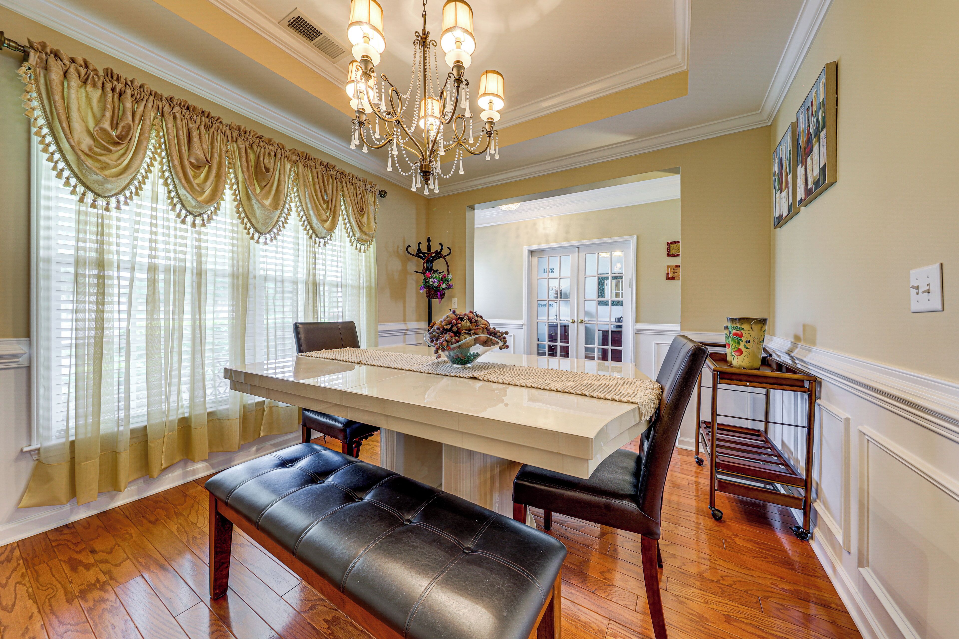 A dining area with a large table, chairs, a bench, a chandelier, and a window with curtains.