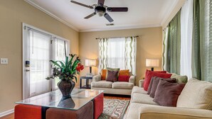 A living room with a glass coffee table, a sofa, a chair, a ceiling fan, and a window with curtains.