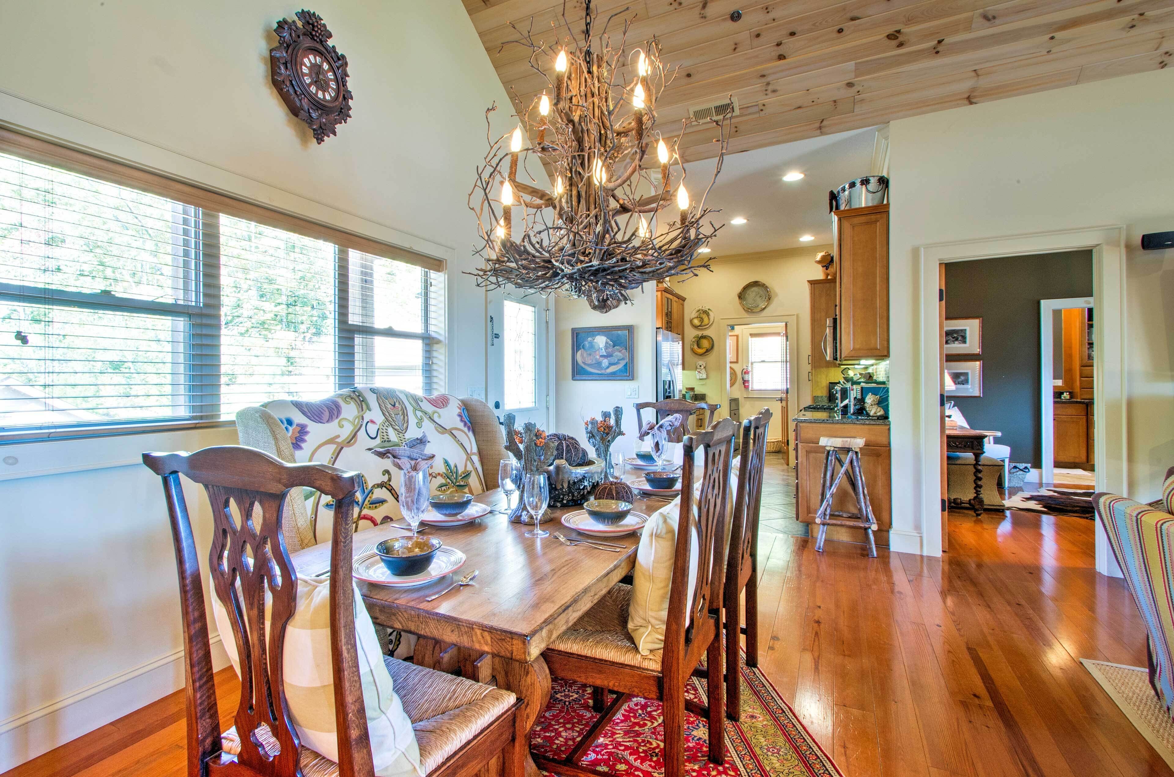 A dining area with a wooden table set for six, surrounded by chairs. A chandelier hangs from the ceiling, and there are windows with blinds on the wall.