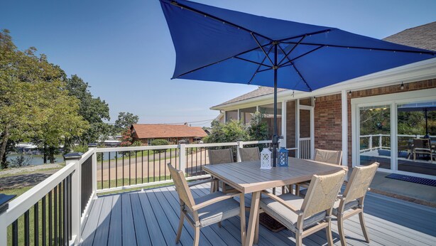 A wooden deck with a table and chairs, a blue umbrella, and a view of a lake and buildings. - Eufaula Oasis w/ Screened Porch: Unwind in Style! (Eufaula)