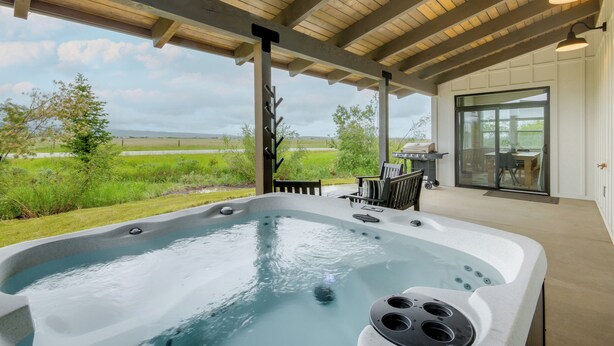 Un bain à remous installé sous une pergola en bois, offrant une vue sur un champ verdoyant et des collines lointaines.
